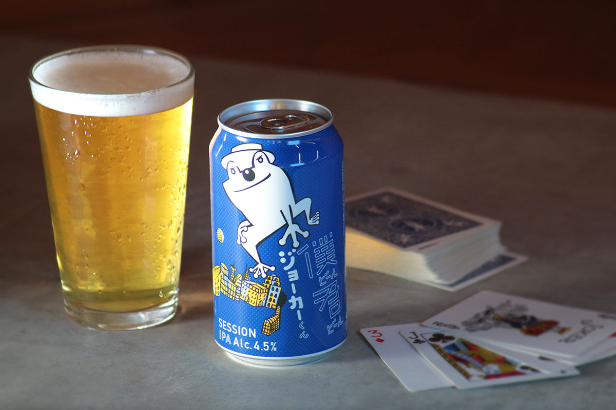 グラスに注いだ僕ビール君ビールジョーカーくんが、缶やトランプと一緒にテーブルの上に並んでいる画像