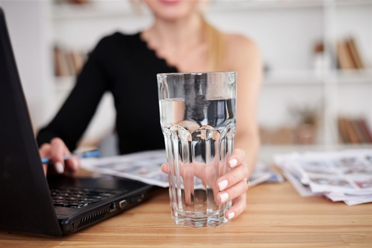 二日酔い対策のため、日中デスクでパソコンを前に働きながらグラスに入った水を飲む女性