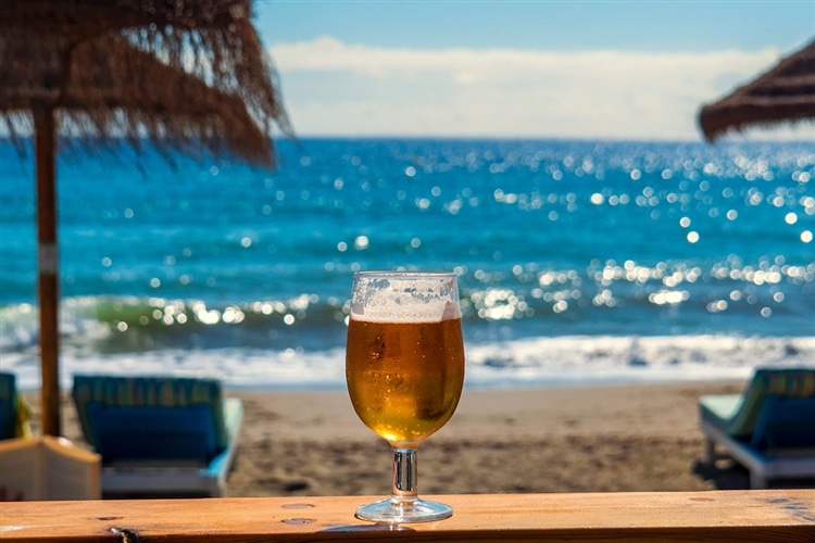 真夏の快晴の海を背景に、手前のテーブルの上に、グラスに注がれたビールが置かれている。置くには青いソファが2つある