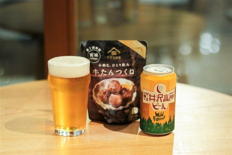 机の上に、軽井沢高原ビール ワイルドフォレストの缶と、ビールが入ったグラス、久世福商店のおつまみ「牛タンつくね」が並んでいる様子