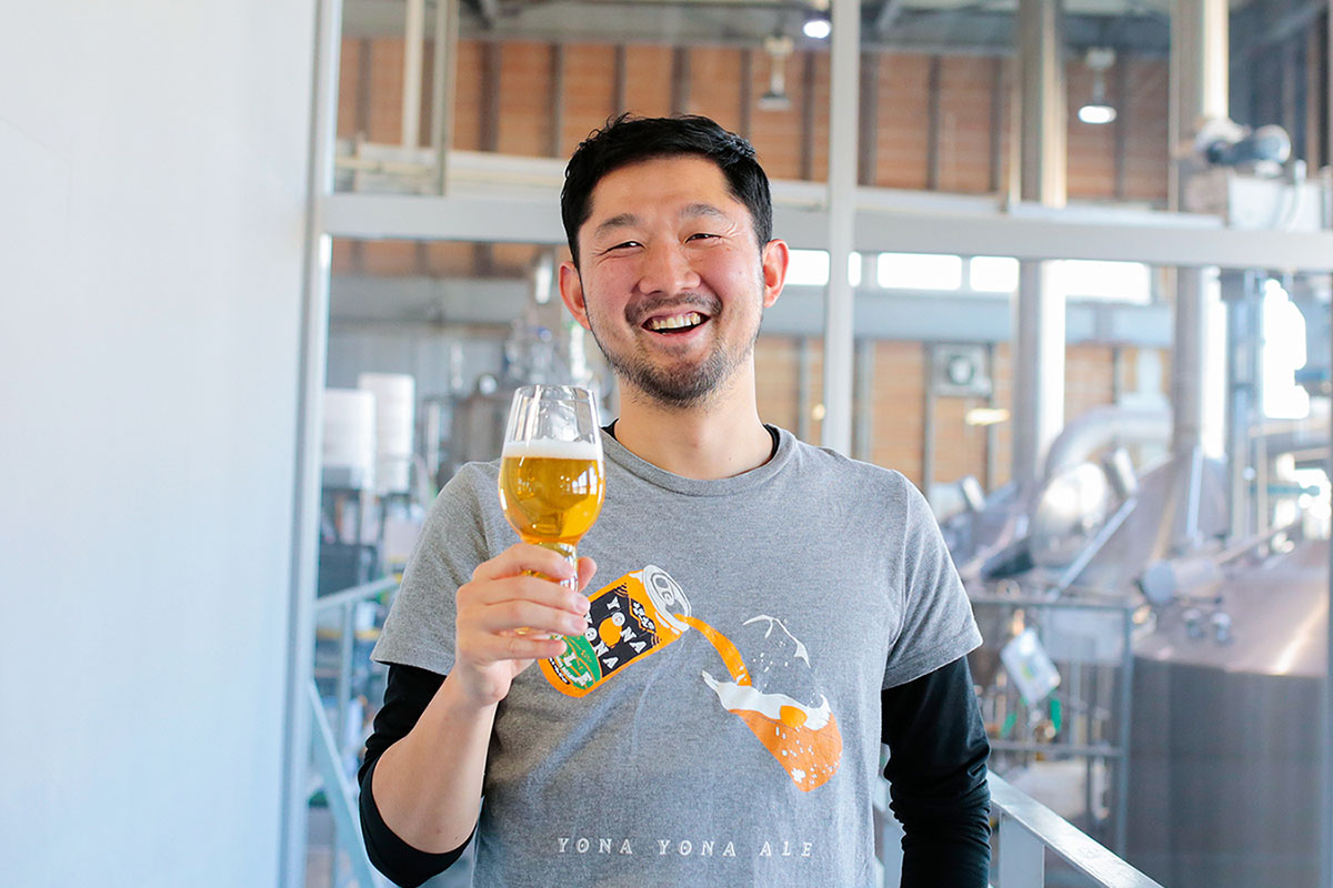 クラフトビールメーカー「ヤッホーブルーイング」 ビール醸造 部門責任者の森田 正文が、醸造所を背景に、ビールが入ったグラスを持って笑っている様子