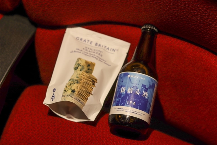 「グレイトブリテン ブルーチーズ クラッカー」とテアトル新宿オリジナルビール「銀幕麦酒」が、映画館の劇場内の椅子の上に載っている様子